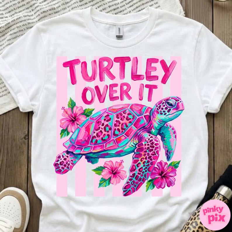 May include: White t-shirt with a colorful graphic of a sea turtle and the text "Turtley Over It" in pink. The turtle has a pink and teal shell with a leopard print design, surrounded by pink flowers.