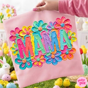 May include: A pink pillow with the word "MAMA" in colorful, textured letters, surrounded by vibrant, multi-colored flowers. The pillow is held up by a person. Easter-themed decorations are in the background.