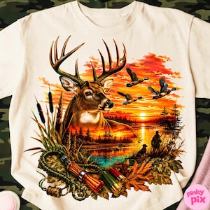 Könnte beinhalten: Ein cremefarbenes T-Shirt mit einem lebendigen Grafikdruck eines Hirschkopfes, fliegenden Enten und einem Sonnenuntergang über einem See. Das Design zeigt Rohrkolben, Herbstblätter, zwei Schrotpatronen sowie einen Jäger mit einem Hund.