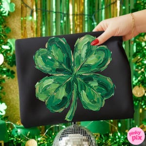 K&ouml;nnte beinhalten: Schwarze Clutch mit einem gro&szlig;en, strukturierten vierbl&auml;ttrigen Kleeblatt-Design in Gr&uuml;n- und Goldt&ouml;nen. Das Kleeblatt ist mit sichtbaren Pinselstrichen gemalt. Die Clutch wird vor einem festlichen gr&uuml;n-goldenen Hintergrund gehalten.