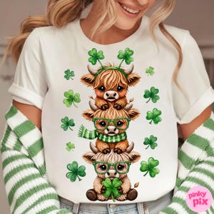 May include: White t-shirt featuring a stack of three cartoon Highland cows, each adorned with St. Patrick's Day accessories. The cows have green glasses, scarves, and shamrock headbands. Green shamrocks are scattered around the design.