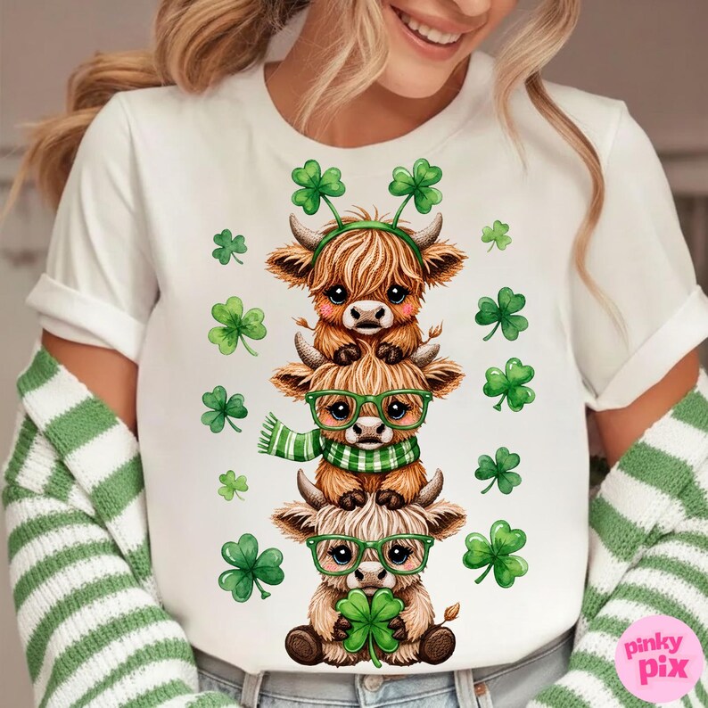 May include: White t-shirt featuring a stack of three cartoon Highland cows, each adorned with St. Patrick's Day accessories. The cows have green glasses, scarves, and shamrock headbands. Green shamrocks are scattered around the design.
