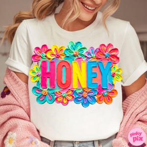 May include: White t-shirt with a colorful floral design and the word "HONEY" in vibrant, multi-colored letters. The design features bright pink, yellow, blue, and green flowers, creating a cheerful and eye-catching look.