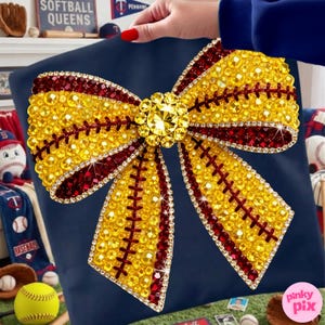 May include: A navy blue pillow featuring a large, embellished bow design. The bow is adorned with yellow and burgundy rhinestones, mimicking a softball. The center of the bow has a large, sparkling gem. The pillow is a decorative item.