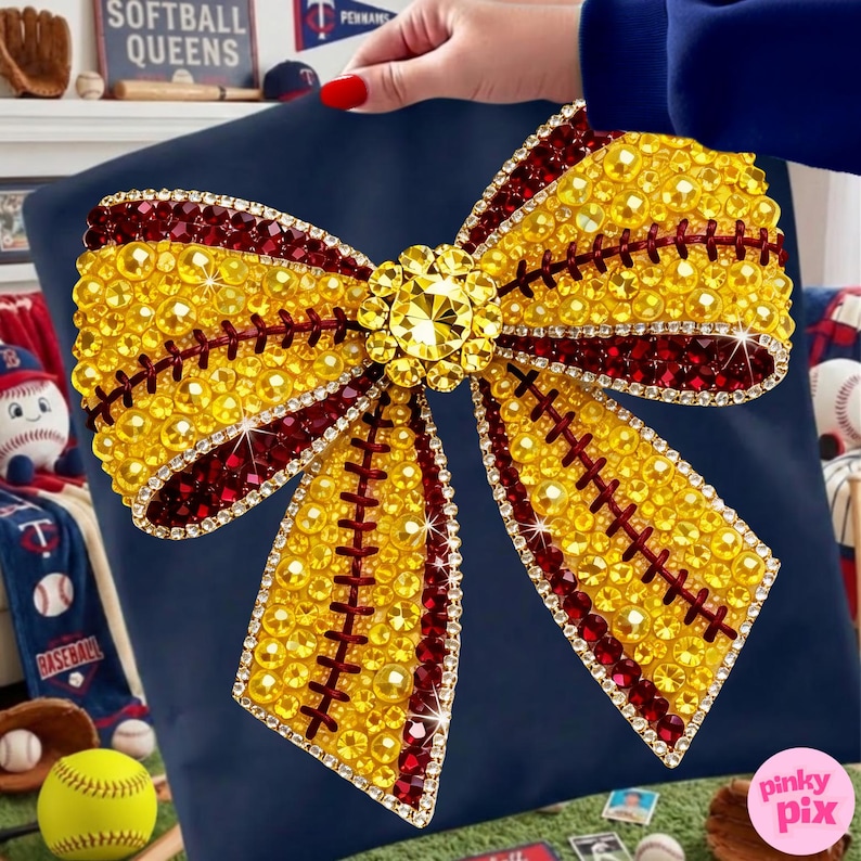May include: A navy cushion with a large, embellished bow design. The bow is decorated with yellow and burgundy rhinestones, resembling a softball. A large, sparkling gem is at the bow's centre. This cushion is a decorative item.