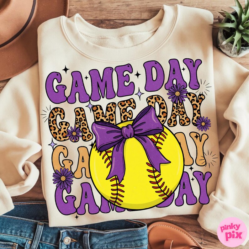 May include: Cream-colored sweatshirt with the text "GAME DAY" in purple, leopard print, and orange lettering. A yellow softball with a purple bow is centered on the front. Purple flowers and starbursts accent the design.