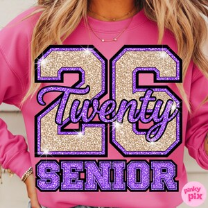 May include: A pink sweatshirt with the text "23 Twenty SENIOR" in a glittery design. The numbers are gold with a purple outline, and the word "Twenty" is in a cursive font. The word "SENIOR" is in purple block letters.