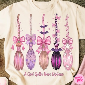 May include: A cream-colored t-shirt featuring five decorative witch brooms in shades of pink and purple. Each broom has a unique design, including bows, bats, and ghosts. The text "A Girl Gotta Have Options" is printed at the bottom.