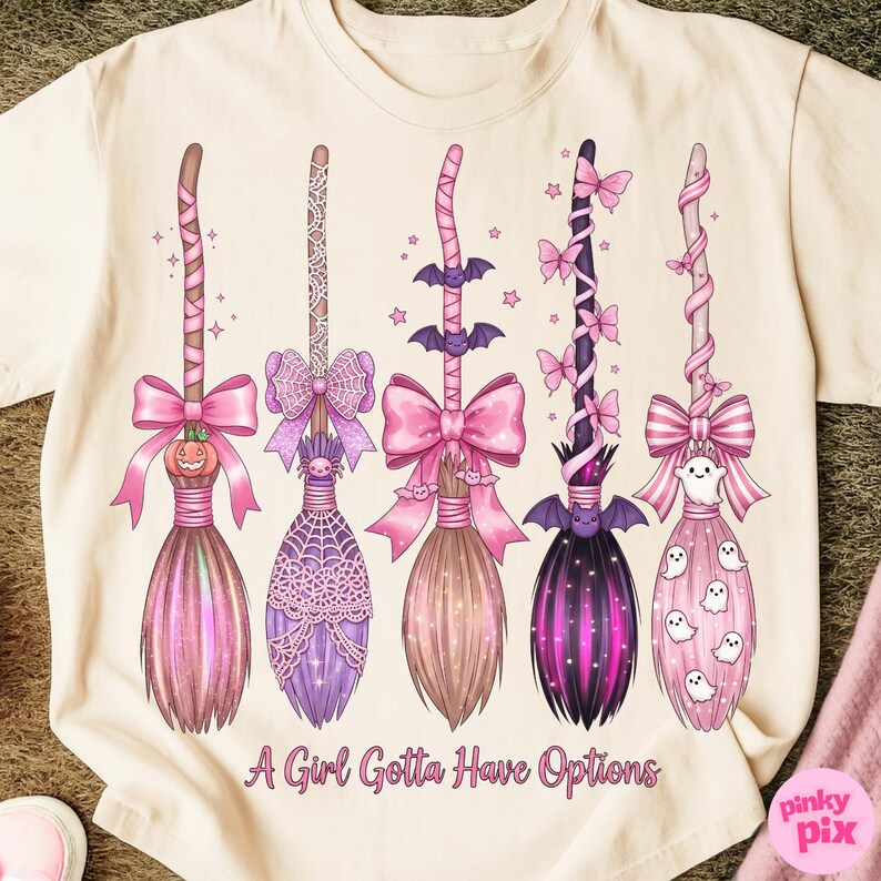 May include: A cream-colored t-shirt featuring five decorative witch brooms in shades of pink and purple. Each broom has a unique design, including bows, bats, and ghosts. The text "A Girl Gotta Have Options" is printed at the bottom.