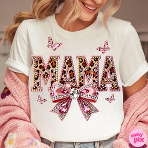 May include: White t-shirt with the word "MAMA" in leopard print letters outlined with rhinestones. A pink bow with a large crystal center and pink rhinestone accents is below the word. Pink butterflies are scattered around the design.