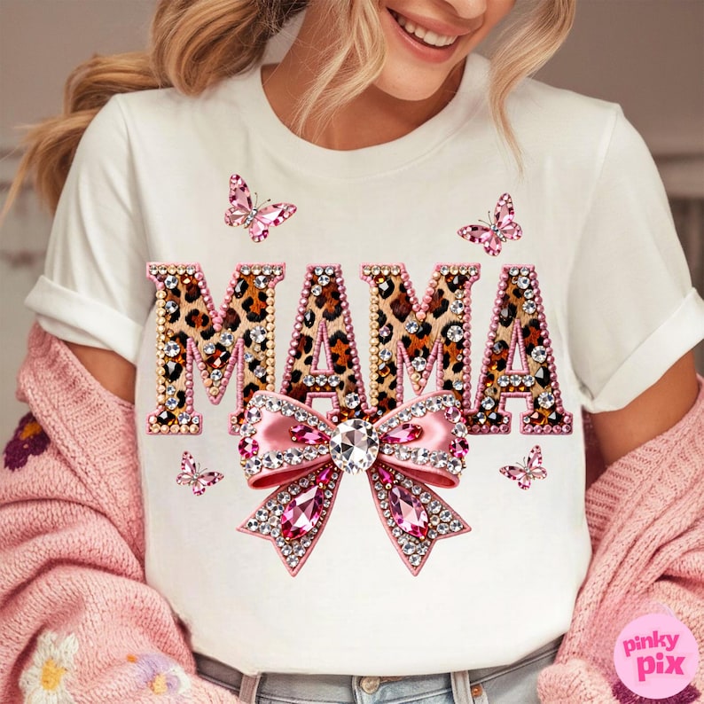 May include: White t-shirt with the word "MAMA" in leopard print letters outlined with rhinestones. A pink bow with a large crystal center and pink rhinestone accents is below the word. Pink butterflies are scattered around the design.