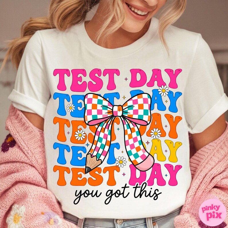 May include: White t-shirt with a colorful graphic that says "TEST DAY" in pink, blue, and orange. A checkered bow and pencil design is in the center. The text "you got this" is at the bottom.