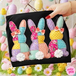 May include: Black t-shirt featuring three colorful patchwork bunny rabbits. Each bunny has a different pattern, including floral, polka dot, and plaid designs. They have peach bows and white fluffy tails. The image is set against a spring-themed background.