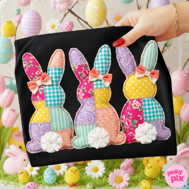 May include: Black t-shirt featuring three colorful patchwork bunny rabbits. Each bunny has a different pattern, including floral, polka dot, and plaid designs. They have peach bows and white fluffy tails. The image is set against a spring-themed background.