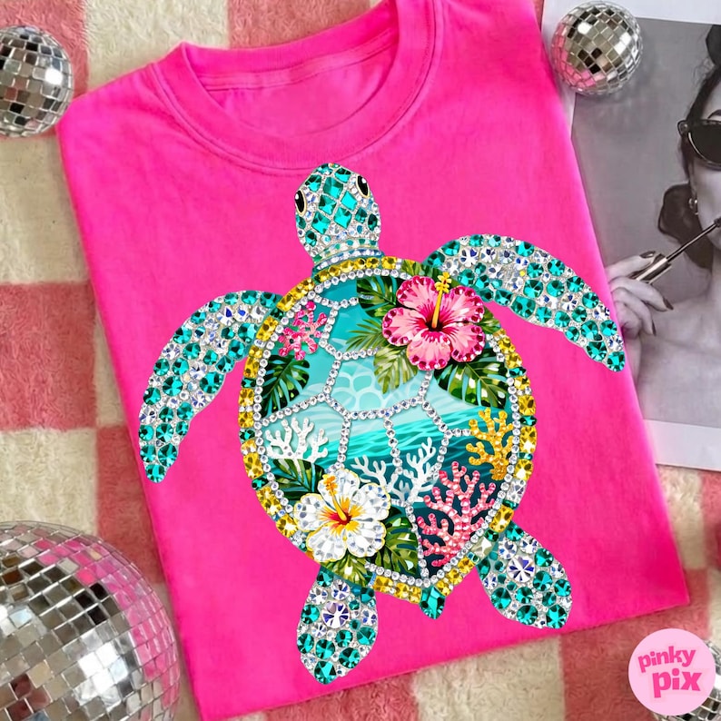 May include: A bright pink t-shirt featuring a bejeweled sea turtle design. The turtle has turquoise, yellow, and clear gem accents, with a turquoise and white ocean scene on its shell. Pink and white hibiscus flowers and coral complete the design.