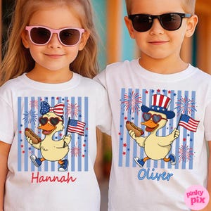 May include: Two children wearing white t-shirts with patriotic duck designs. Each shirt features a cartoon duck with sunglasses, holding a hot dog and an American flag. The shirts have blue and white striped backgrounds, fireworks, and the names "Hannah" and "Oliver".