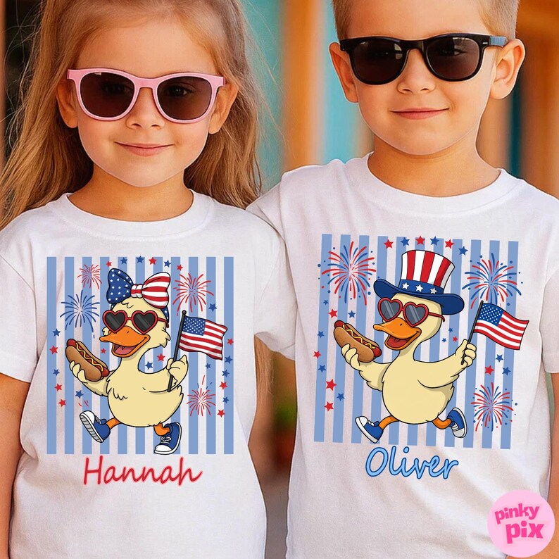 May include: Two children wearing white t-shirts with patriotic duck designs. Each shirt features a cartoon duck with sunglasses, holding a hot dog and an American flag. The shirts have blue and white striped backgrounds, fireworks, and the names "Hannah" and "Oliver".