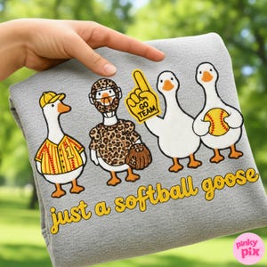 May include: A gray, soft fabric item featuring four cartoon geese dressed in softball gear. One goose wears a baseball uniform, another a leopard-print outfit, and a third holds a foam finger. The text "just a softball goose" is in yellow.