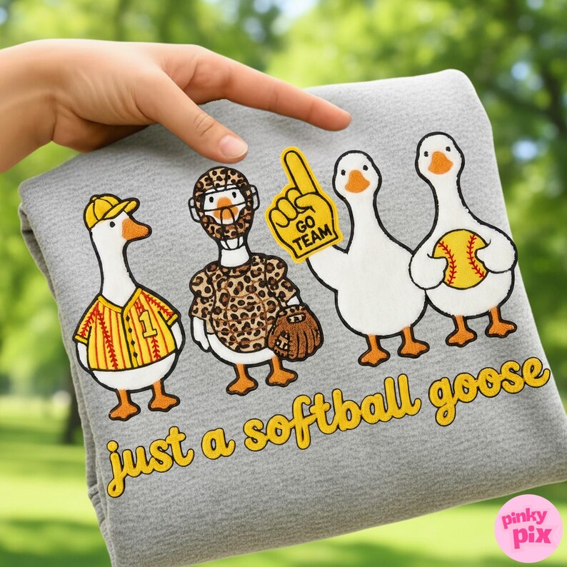 May include: A gray, soft fabric item featuring four cartoon geese dressed in softball gear. One goose wears a baseball uniform, another a leopard-print outfit, and a third holds a foam finger. The text "just a softball goose" is in yellow.