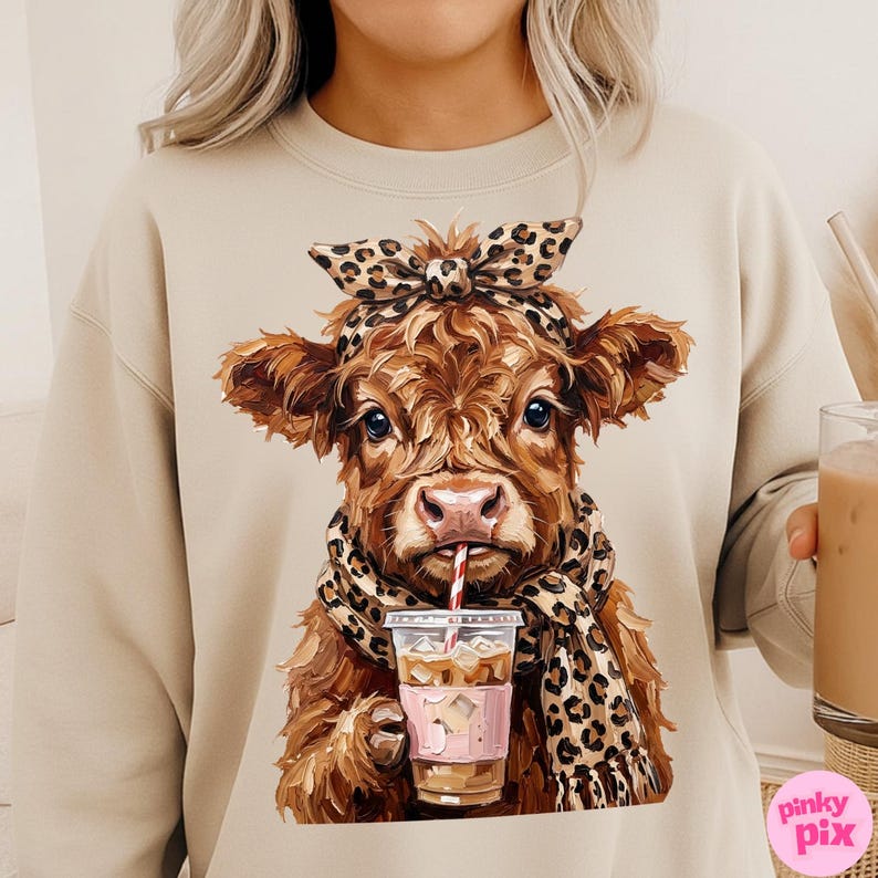 May include: Beige sweatshirt featuring a watercolor illustration of a brown cow wearing a leopard print scarf and headband. The cow is drinking from a coffee cup with a red and white striped straw. The sweatshirt has a crew neck.