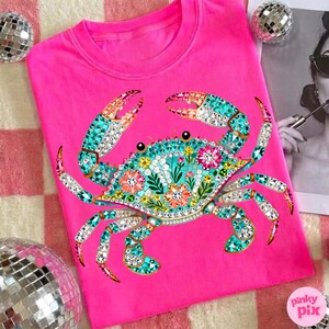 May include: A bright pink t-shirt featuring a detailed crab design. The crab is adorned with turquoise, crystal, and floral accents. The shirt has a crew neck and short sleeves. The Pinky Pix logo is visible in the bottom right corner.