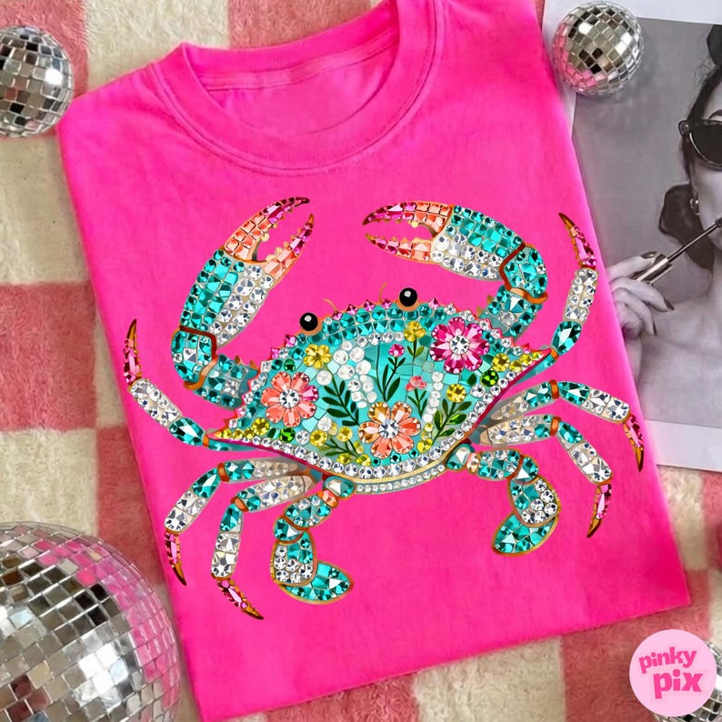 May include: A bright pink t-shirt featuring a detailed crab design. The crab is adorned with turquoise, crystal, and floral accents. The shirt has a crew neck and short sleeves. The Pinky Pix logo is visible in the bottom right corner.