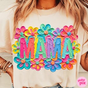 May include: A cream-colored t-shirt with the word "MAMA" in colorful, textured letters, surrounded by vibrant, 3D-effect flowers in shades of pink, yellow, blue, and orange. The shirt has short sleeves.