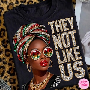 May include: Black t-shirt featuring a vibrant illustration of a woman wearing a jeweled head wrap, sunglasses, and jewelry. The text "THEY NOT LIKE US" is displayed in jeweled letters. The image has a leopard print background.
