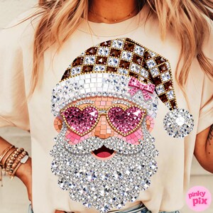 May include: A beige t-shirt featuring a graphic of a Santa Claus face composed of sparkling rhinestones. The design includes a brown and white checkered hat with a pink bow, heart-shaped pink sunglasses, and a large, textured white beard.