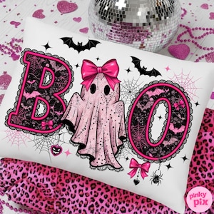 Puede incluir: Coj&iacute;n rectangular blanco con tem&aacute;tica de Halloween. La palabra "BOO" est&aacute; escrita con letras de encaje rosa y negro. Un fantasma rosa con un lazo est&aacute; en el centro. Murci&eacute;lagos, telara&ntilde;as y lazos decoran el coj&iacute;n. Una bola de discoteca y tela con estampado de leopardo rosa est&aacute;n en el fondo.