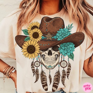 Op de afbeelding: Cr&egrave;mekleurig T-shirt met een gedetailleerde afbeelding van een schedel met een bruine cowboyhoed versierd met zonnebloemen, turquoise bloemen en een ster. Dromenvangers en veren hangen eronder. Western- en bohemien thema.