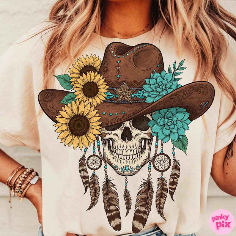 Op de afbeelding: Cr&egrave;mekleurig T-shirt met een gedetailleerde afbeelding van een schedel met een bruine cowboyhoed versierd met zonnebloemen, turquoise bloemen en een ster. Dromenvangers en veren hangen eronder. Western- en bohemien thema.