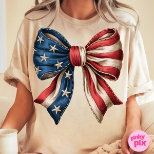 Puede incluir: Una camiseta beige con un gran lazo rojo, blanco y azul impreso en la parte delantera. El lazo est&aacute; dise&ntilde;ado para parecerse a la bandera estadounidense.