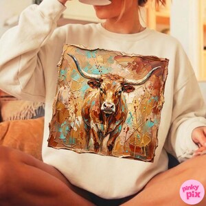 May include: A cream-colored sweatshirt featuring a detailed painting of a longhorn steer. The artwork uses a palette of browns, tans, and turquoise, with visible brushstrokes. The steer has large horns and a focused gaze, creating a rustic, artistic aesthetic.