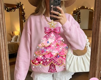 Może przedstawiać: Jasnoróżowa bluza z nadrukiem choinki. Choinka ozdobiona jest różowymi i czerwonymi klejnotami, gwiazdą i kokardą. Bluza jest połączona z białymi szortami i słomkowym kapeluszem. Biała torebka trzymana jest w prawej dłoni.