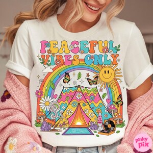 May include: White t-shirt with a colorful graphic design. The design features a rainbow, butterflies, a smiling sun, flowers, mushrooms, and a tent with the text "PEACEFUL VIBES ONLY". A retro-style lava lamp and record player are also included.