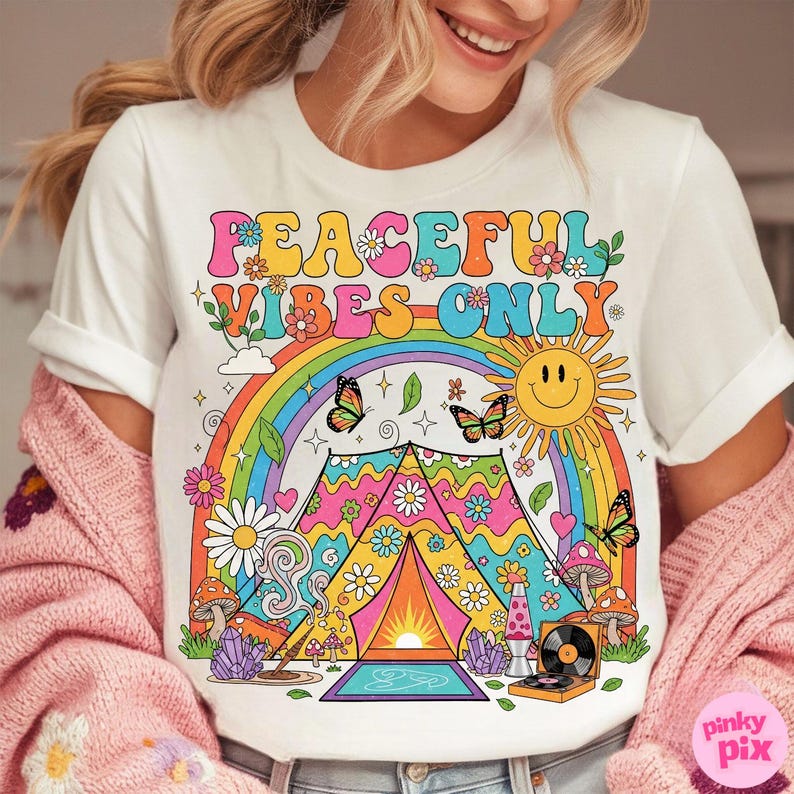May include: White t-shirt with a colorful graphic design. The design features a rainbow, butterflies, a smiling sun, flowers, mushrooms, and a tent with the text "PEACEFUL VIBES ONLY". A retro-style lava lamp and record player are also included.