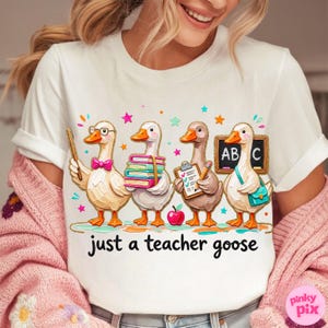 May include: White t-shirt featuring a whimsical illustration of four geese in a classroom setting. The geese are depicted with school supplies and the text "just a teacher goose". Colorful stars and an apple are also included in the design.