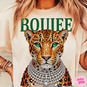May include: Cream-colored t-shirt featuring a jeweled leopard design with green eyes and a diamond necklace. The word "BOUIEE" is printed in green above the leopard. The shirt is a casual wear item.