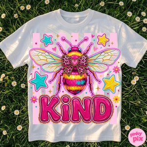 May include: White t-shirt featuring a colorful bee design with a bow and the word "KIND" in pink. The bee has yellow, pink, and black stripes. The design includes stars and flowers on a pink background.