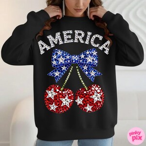 May include: Black sweatshirt with the word "AMERICA" in white, sparkling letters. A blue bow with white stars sits above two red cherries with white stars. The cherries have green stems. The design is centered on the front of the sweatshirt.