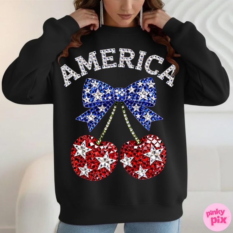 May include: Black sweatshirt with the word "AMERICA" in white, sparkling letters. A blue bow with white stars sits above two red cherries with white stars. The cherries have green stems. The design is centered on the front of the sweatshirt.