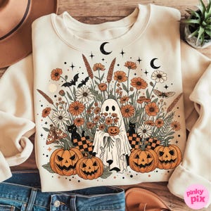 May include: Cream-coloured sweatshirt with a Halloween graphic. The design shows a ghost holding a pumpkin, surrounded by orange flowers, pumpkins, black cats, bats, and crescent moons. The graphic has a vintage, whimsical style.