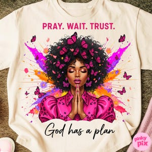 May include: Cream-colored t-shirt featuring a vibrant illustration of a woman with closed eyes, adorned with pink butterflies in her hair and earrings. The design includes the text "PRAY. WAIT. TRUST." and "God has a plan."