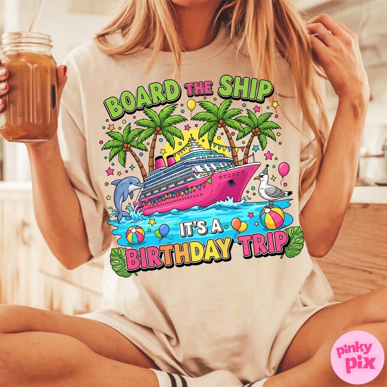 May include: Beige t-shirt featuring a vibrant graphic of a pink cruise ship, palm trees, and the text "BOARD THE SHIP IT'S A BIRTHDAY TRIP". The design includes a dolphin, seagull, and beach balls.