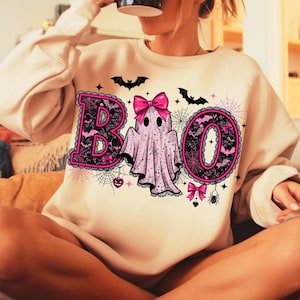 Puede incluir: Sudadera beige con un dise&ntilde;o de Halloween. La palabra "BOO" est&aacute; escrita en rosa y negro, con un fantasma en el centro. Murci&eacute;lagos, telara&ntilde;as y lazos decoran el dise&ntilde;o, creando un aspecto festivo.