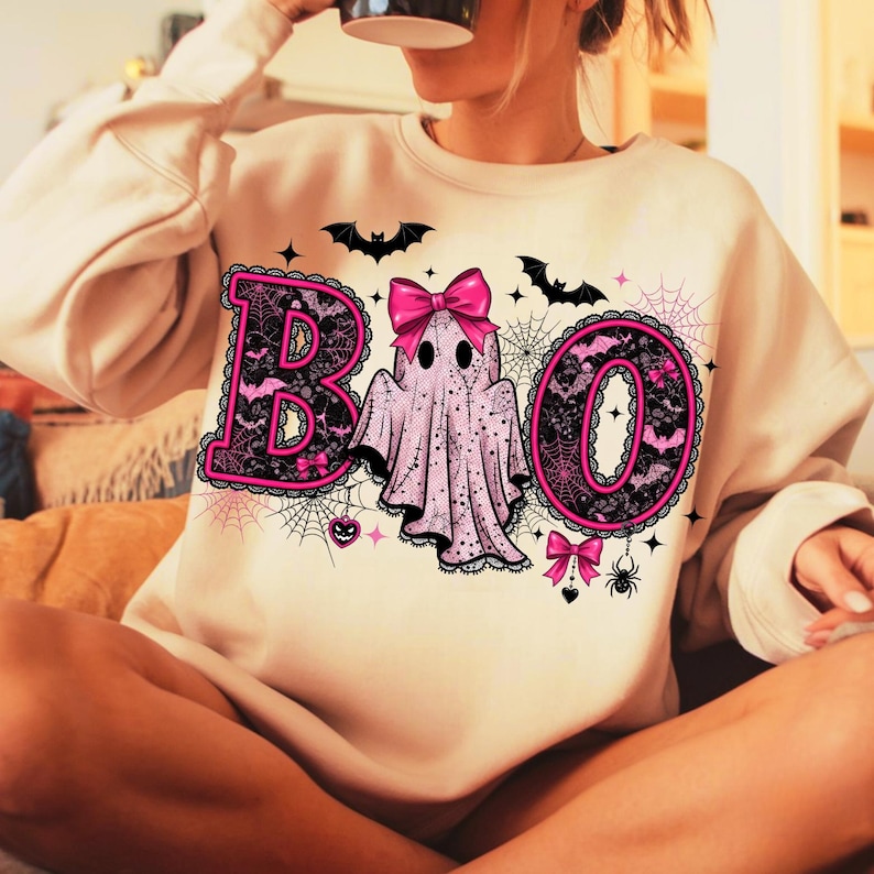 Puede incluir: Sudadera beige con un dise&ntilde;o de Halloween. La palabra "BOO" est&aacute; escrita en rosa y negro, con un fantasma en el centro. Murci&eacute;lagos, telara&ntilde;as y lazos decoran el dise&ntilde;o, creando un aspecto festivo.