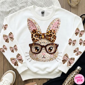 May include: White sweatshirt with a bedazzled bunny face wearing glasses and a leopard print bow. The sleeves are decorated with leopard print and pink bow accents. A fun and festive Easter-themed top.