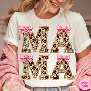 May include: White t-shirt featuring the word "MAMA" in a leopard print pattern, adorned with rhinestones and pink bows. A casual top suitable for everyday wear.