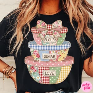May include: Black t-shirt featuring a stack of patchwork bowls with the words "FLOUR", "SUGAR", and "LOVE". The bowls have floral, polka dot, and gingham patterns. A large bow sits atop the top bowl.